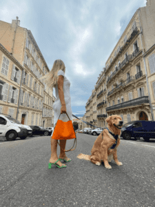 Au centre d'une route sereine, une femme, son sac en cuir orange à la main, est accompagnée de son fidèle chien, tous deux capturés dans un moment de tranquillité inattendue.