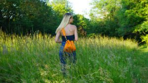 Une femme blonde, élégante, se tient au milieu d'une forêt luxuriante, son sac en cuir orange contrastant magnifiquement avec les teintes verdoyantes environnantes.
