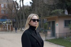 Portrait d'une femme élégante portant des lunettes de soleil noires et un manteau noir, se tenant dans un parc urbain.